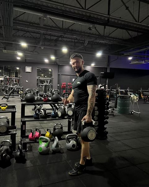 Mateusz z hantlem w dłoni na siłowni, obok stojak z kettlebellami